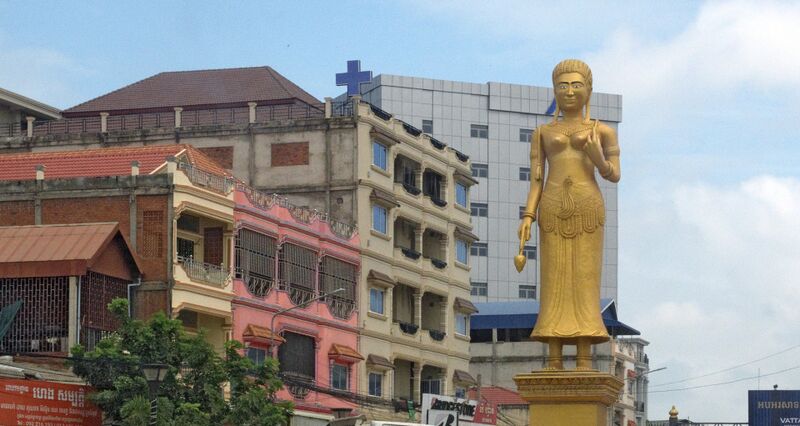 A tall gilded statue of a standing woman in a long skirt rises on a gold pedestal beside a row of shophouses, one painted pink, with a taller office building behind under a pale blue sky.
