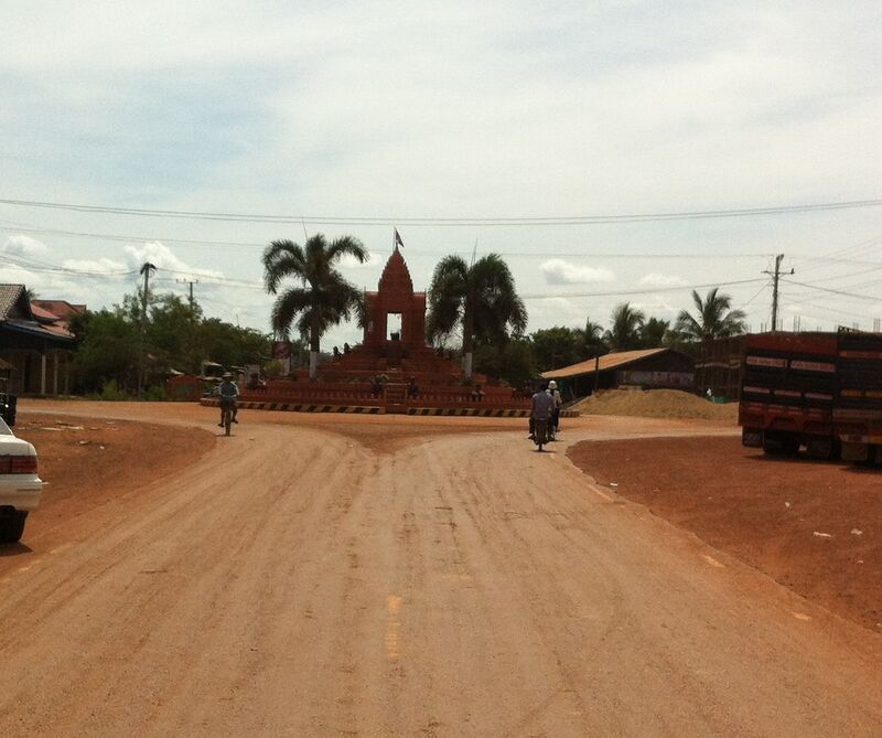A wide red dirt road leads to a roundabout where a red temple-style tower with a flag on top rises from a stepped base, between two palm trees. Two motorbikes ride toward it, a sand pile and a large truck stand at the right, and low buildings and trees line the edges under a cloudy sky.
