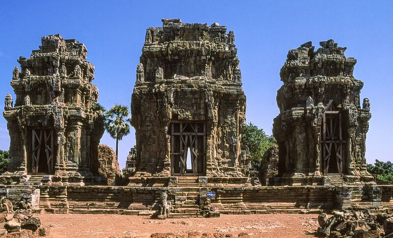 Three weathered sandstone temple towers in a row on a hilltop, under a deep blue sky, with a sugar palm between two of them.