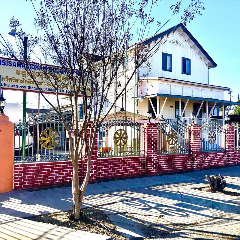 A white two-story wooden building behind a red brick fence with gold Dharma wheels, a bare tree and a temple sign in front.