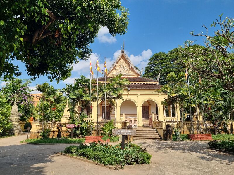 A cream temple hall with arched porch openings and a pointed gable, behind a garden and a curving path.