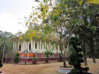 A long white temple hall with an orange roof among trees, with clipped shrubs in round planters in front.