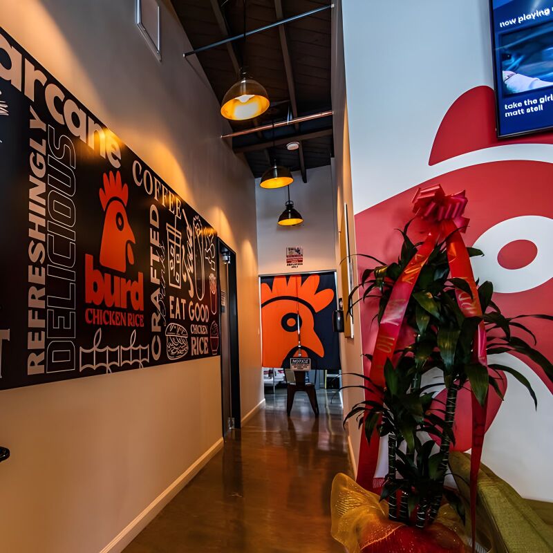 A narrow restaurant hallway with a black mural wall of slogans, a red rooster logo and a potted plant.
