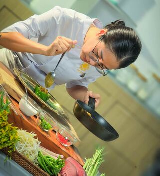 A chef in a white jacket leans over a pan, spooning sauce, with trays of fresh vegetables in front.