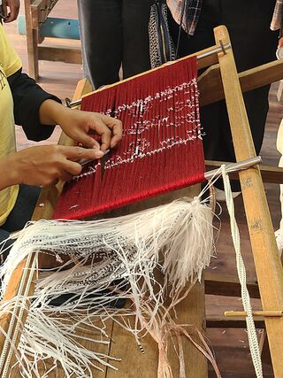 Hands pulling plastic ties from bundles of red-dyed silk threads stretched on a wooden frame.