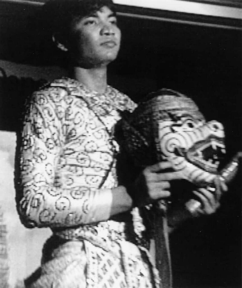A black-and-white photo of a young man in a patterned costume looking to one side, holding a large carved Hanuman mask.