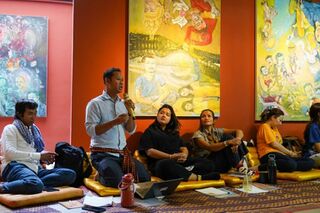 Adults sitting on cushions on the floor in a room with large colorful paintings, listening to a speaker.