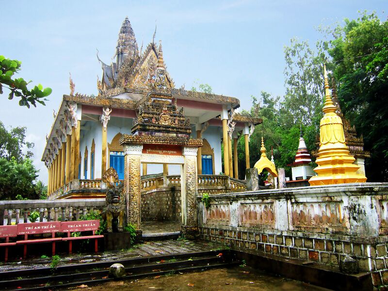 A Khmer temple with a tall tiered spire behind a wall and a pond, with a gilded stupa at the right.