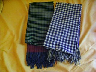 Two folded cotton scarves, one green and one blue, in a checked pattern with fringes, on a yellow cloth.