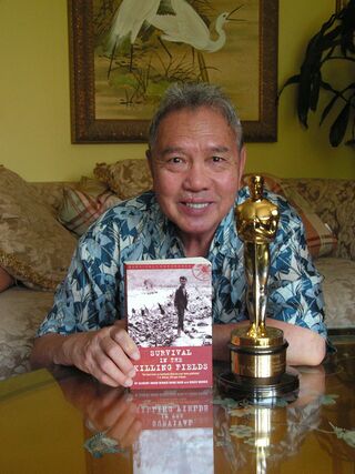 A man at a table holds up a book beside a gold Oscar statuette.
