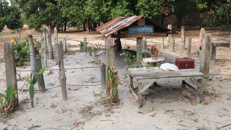 A bare sandy clearing with low wooden posts and a rope fence around a plain wooden table and a small tin-roofed shelter, with trees behind.