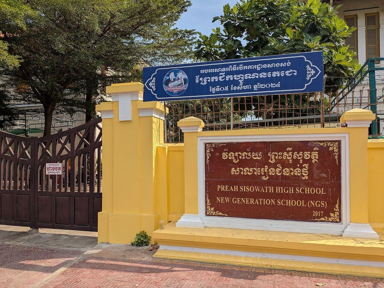 A yellow school gate and wall with a blue name sign and a red plaque in Khmer and English, under trees